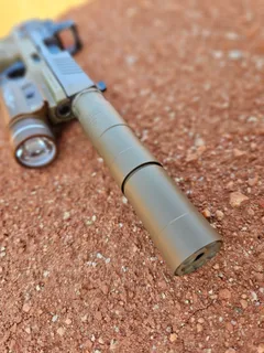 Rugged Oculus 22 suppressor in flat dark earth mounted on a pistol, lying on red dirt, viewed toward the muzzle